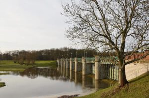 Presa de pilares y compuertas junto a orilla arbolada reflejada en el agua, mostrando agua y tecnologia en gestión de caudal.