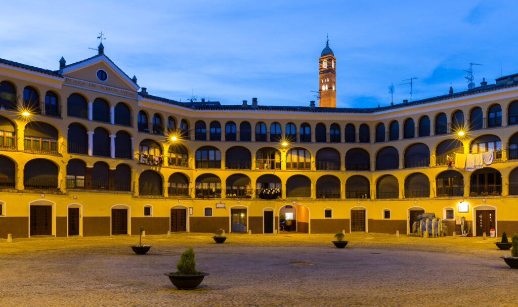 Plaza porticada iluminada al atardecer con fachada coral restaurado y torre campanario