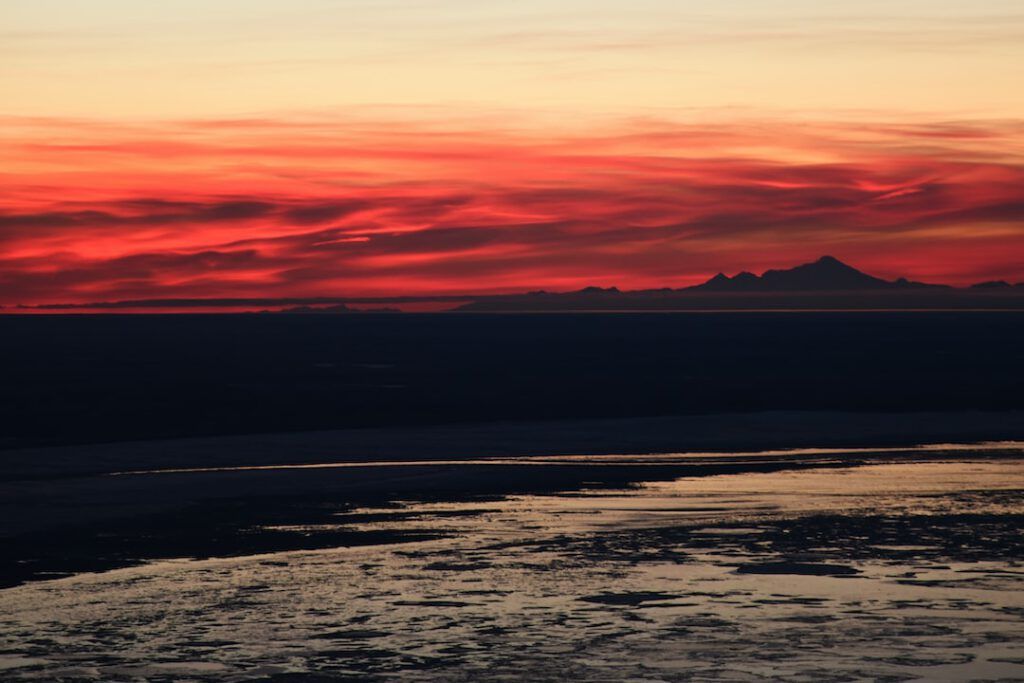 Atardecer rojo sobre el mar con reflejos en la superficie y silueta de montañas en el horizonte, costos vehiculo electrico vs gasolina descubre verdad aqui