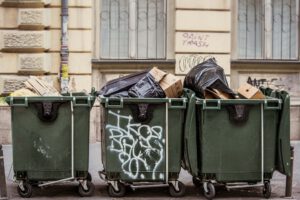 Tres contenedores verdes llenos de bolsas y cartón junto a una fachada, metales reciclados 1.