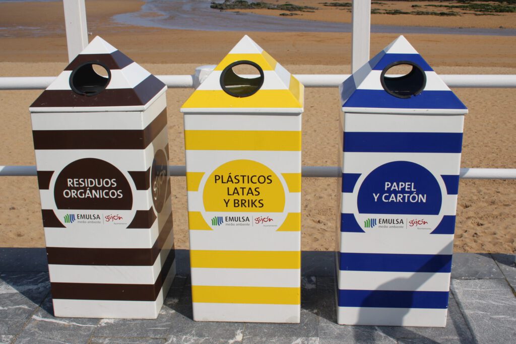 Tres contenedores rayados en la playa destinados al reciclaje de plasticos 3, papel y residuos orgánicos.
