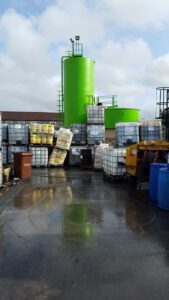Tanques cilíndricos verdes y contenedores IBC apilados en un área exterior mostrando medidas de seguridad quimica